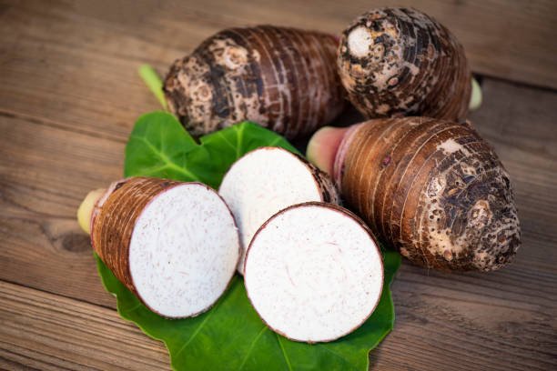 A bowl of cooked taro chunks showing its purple and white flesh, with some raw taro roots beside it.